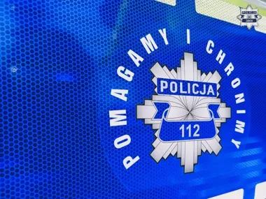 Fragment niebieskiego munduru lub pojazdu policji z napisem „POMagamy i chronimy”, emblematem POLICJA oraz numerem 112, w prawym górnym rogu widoczny znak komen