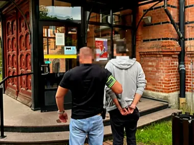 Funkcjonariusz w czarnej koszulce z opaską na ramieniu prowadzi w kajdankach mężczyznę w szarej bluzie przed wejściem do ceglanego budynku.