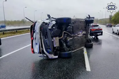 Przewrócony samochód osobowy leży na boku na mokrej autostradzie, obok stoi uszkodzony czarny hatchback; w tle jadą inne pojazdy.