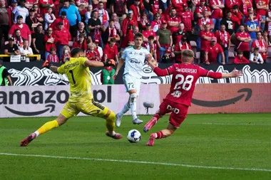 Piłkarz w czerwonym stroju z numerem 28 strzela do bramki, a bramkarz w żółtym stroju z numerem 1 próbuje obronić strzał podczas meczu ligowego na stadionie z w