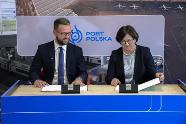 Mężczyzna i kobieta w garniturach siedzą przy niebieskim stole i podpisują dokumenty podczas uroczystości z logo Port Polska na tle wizualizacji lotniska.